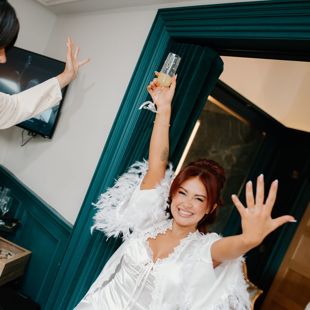 Bride in a feather-trimmed satin robe raises a glass and shows off her ring during morning prep