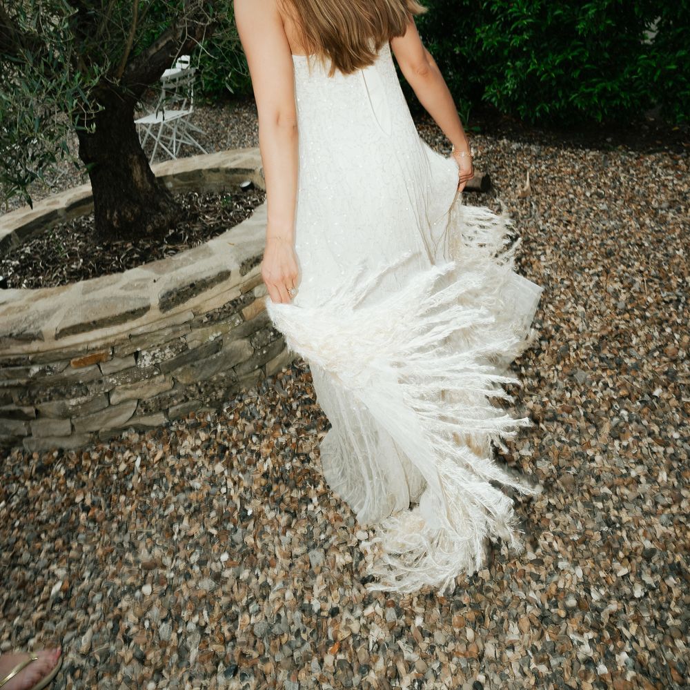 Bride lifting the fringed hem of her strapless gown beside an olive tree and gravel path