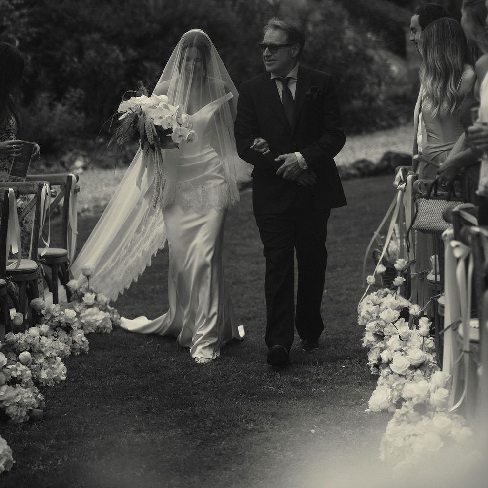 Bride in a lace-trimmed veil walks down a floral-lined garden aisle on a man's arm.