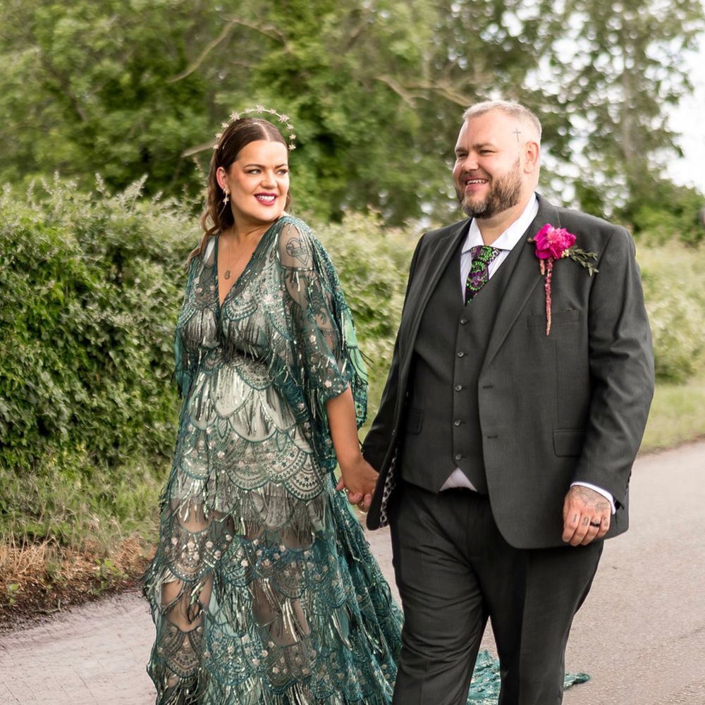 Bride in a green fringed gown and groom in a grey suit walking hand in hand down a country lane