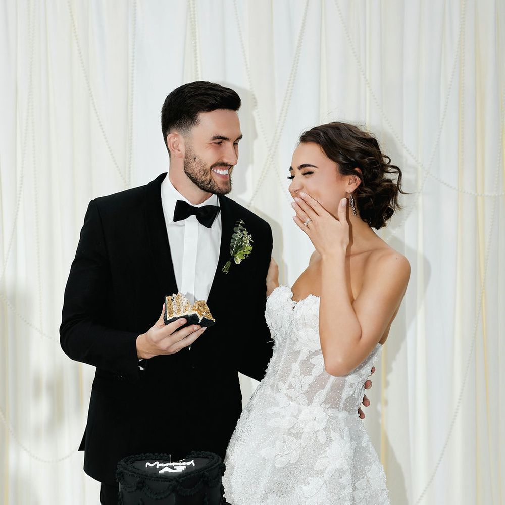 Laughing bride in a short lace dress and groom in black tuxedo beside a black wedding cake
