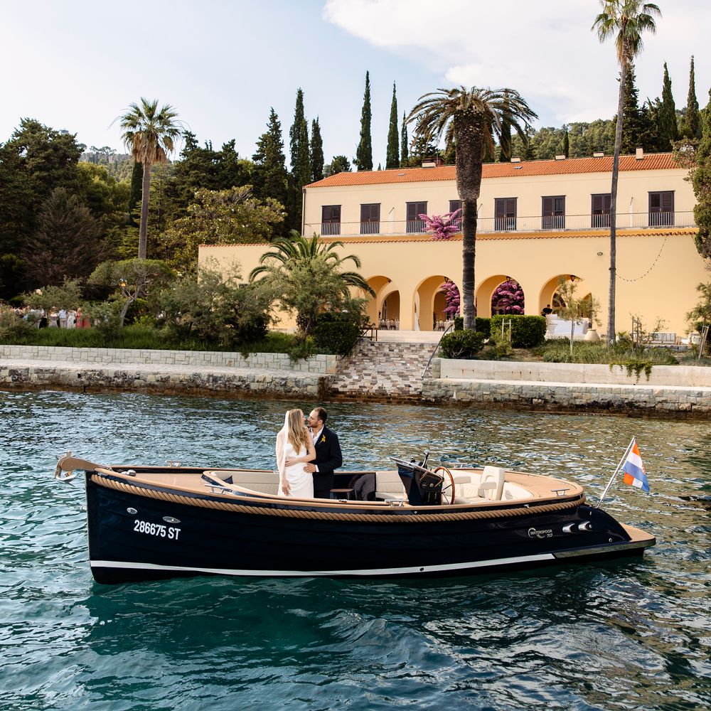 Bride and groom embrace aboard a wooden boat on turquoise water before a Mediterranean villa