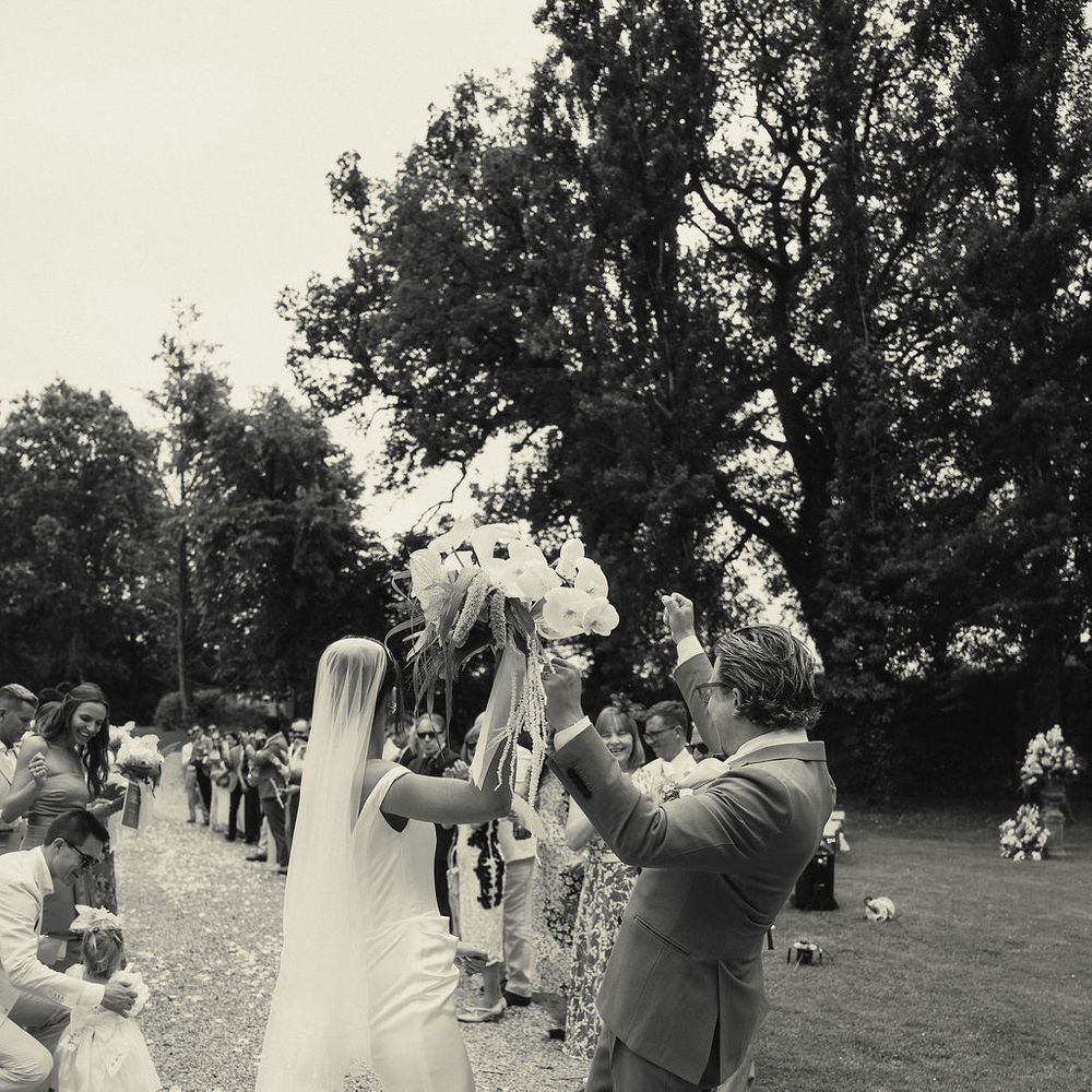 Bride and groom raise her bouquet high as guests celebrate on a tree-lined lawn