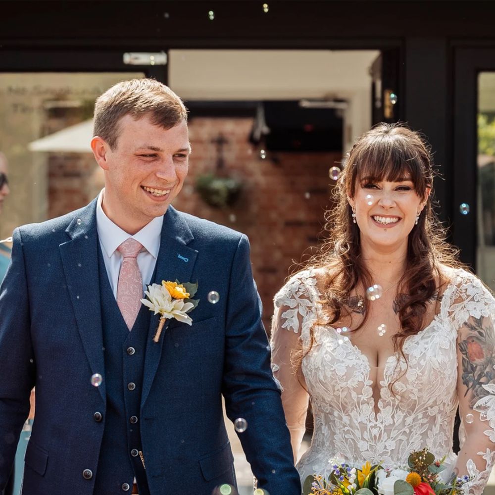 Smiling bride and groom hold hands amid bubbles, she carries a bright sunflower bouquet