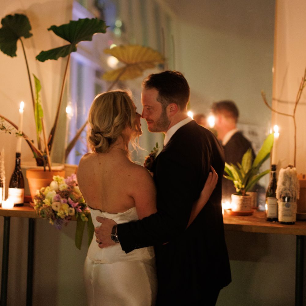 Bride and groom embrace by candlelight surrounded by potted plants and taper candles