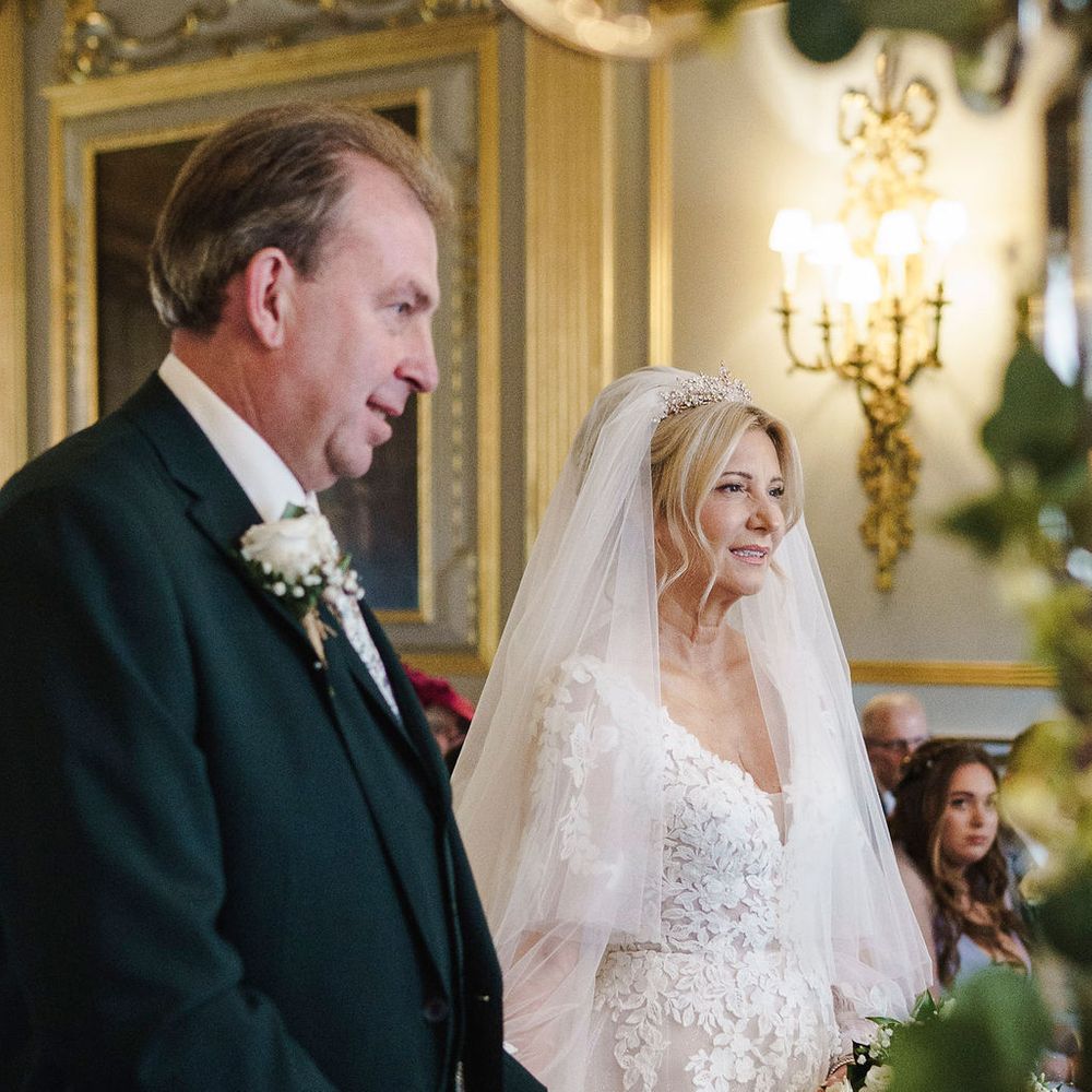 Bride in lace gown and tiara stands beside groom during an elegant indoor ceremony