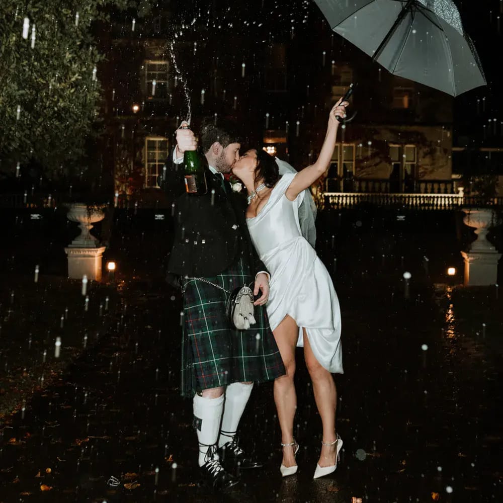 Groom in kilt and bride sharing a kiss, spraying champagne under an umbrella in the rain at night