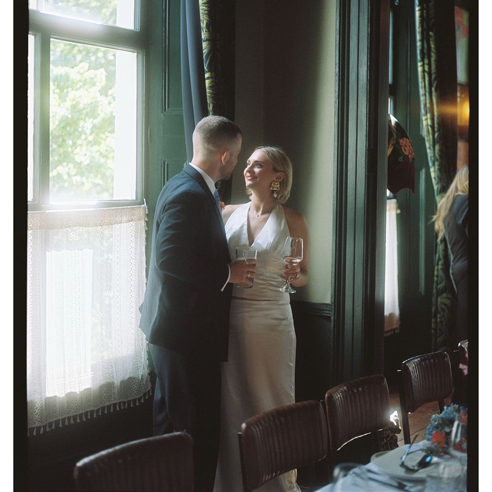 Bride and groom sharing a quiet moment with champagne by a lace-curtained window