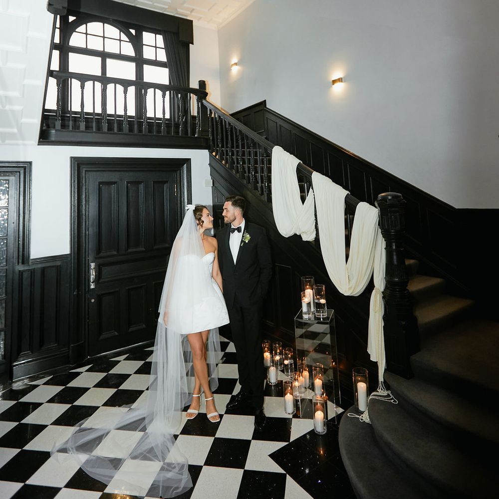 Bride and groom stand beneath a crystal chandelier on a black and white chequered floor by a dark staircase