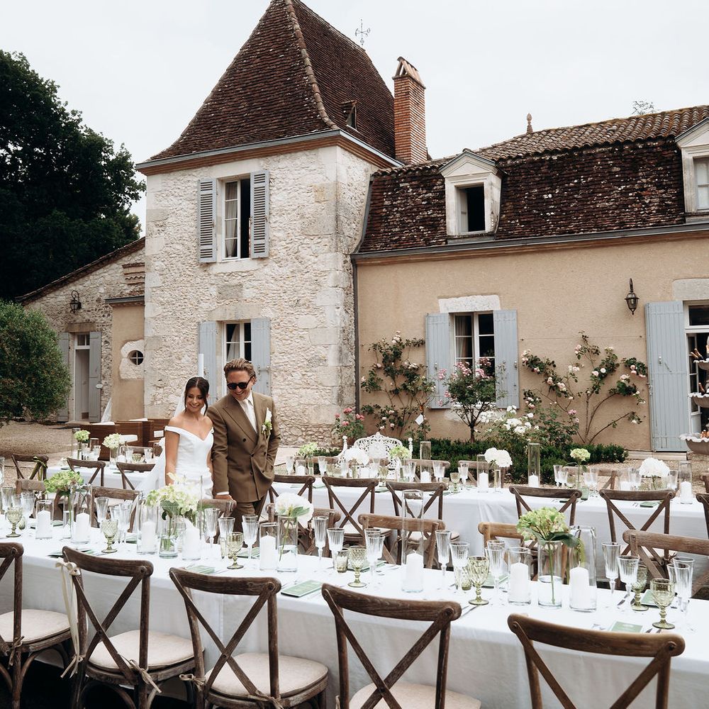 Bride and groom stand beside long banquet tables outside a French chateau with blue shutters