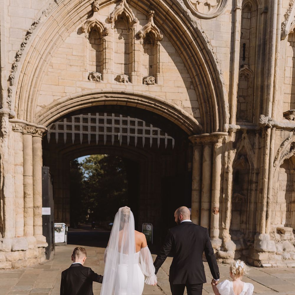 Bride and groom walk hand in hand with two children towards a Gothic stone archway.
