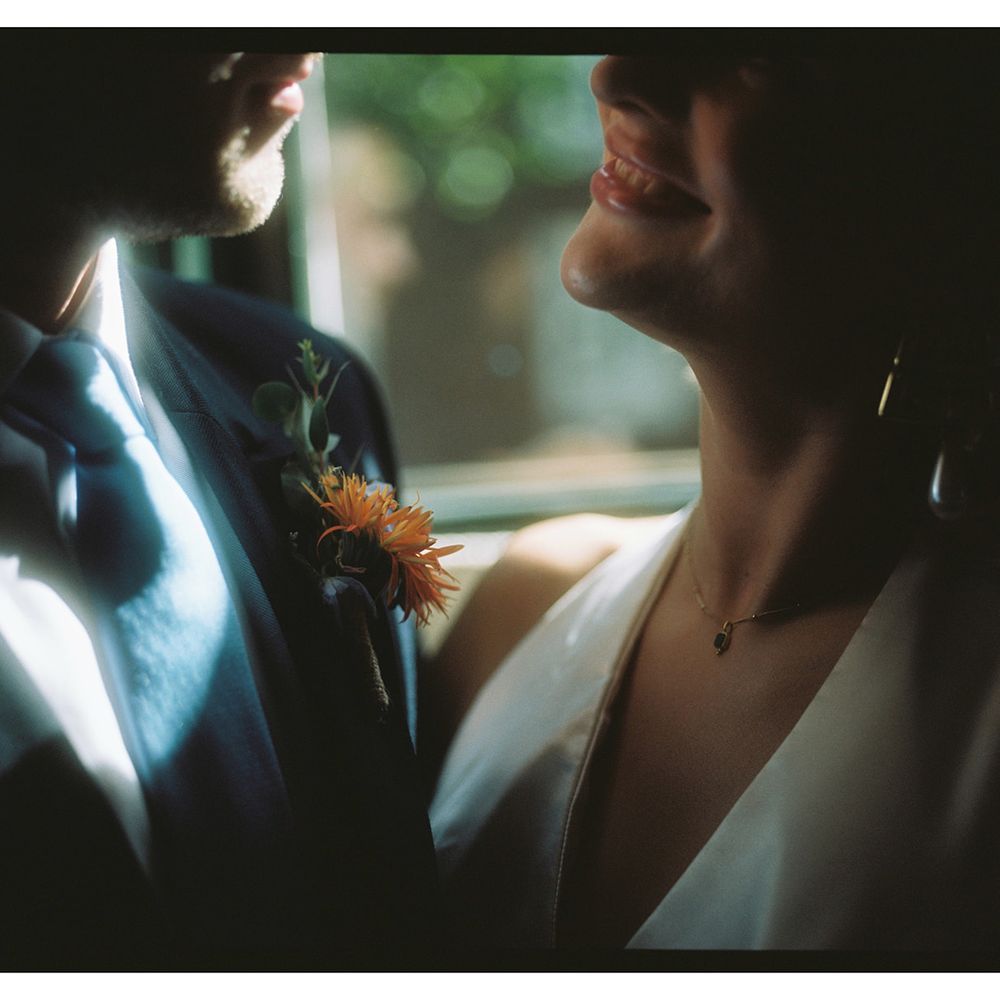 Groom in navy suit with orange buttonhole stands close to smiling bride in plunging dress