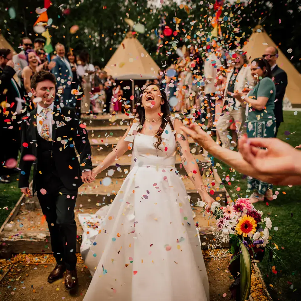 Laughing bride and groom walk through a shower of colourful confetti as guests cheer outside tipis