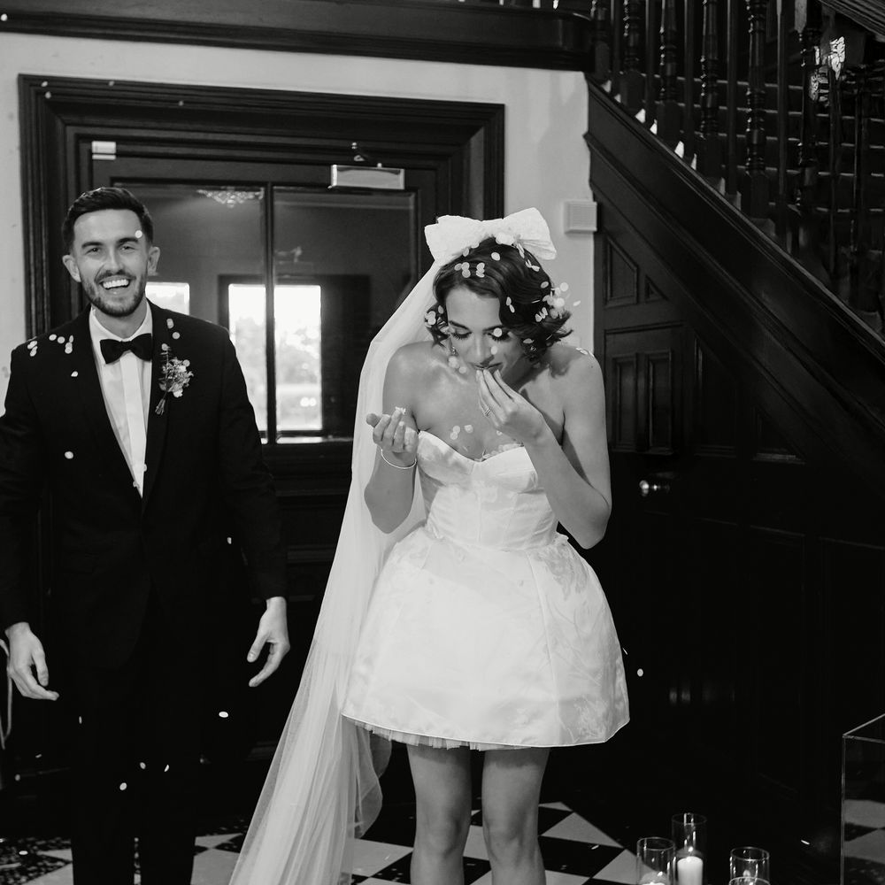 Laughing bride in short dress and long veil beside groom on chequered floor with candles