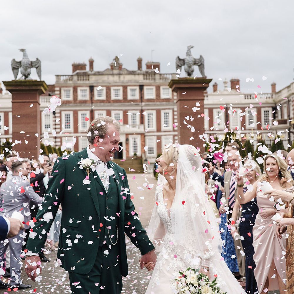 Bride and groom hold hands walking through pink and white confetti outside a grand country house
