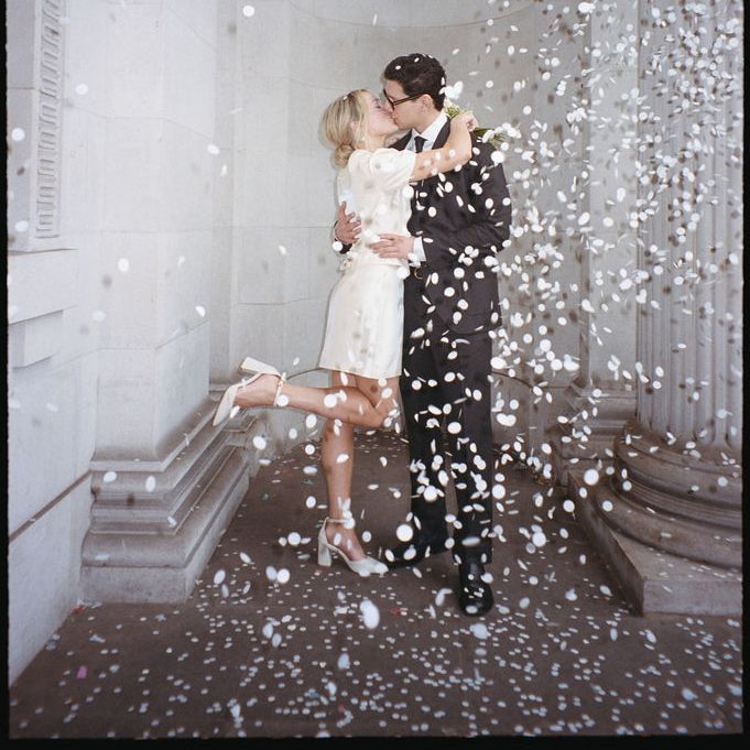 Bride in short dress and groom kissing amid falling confetti between stone pillars