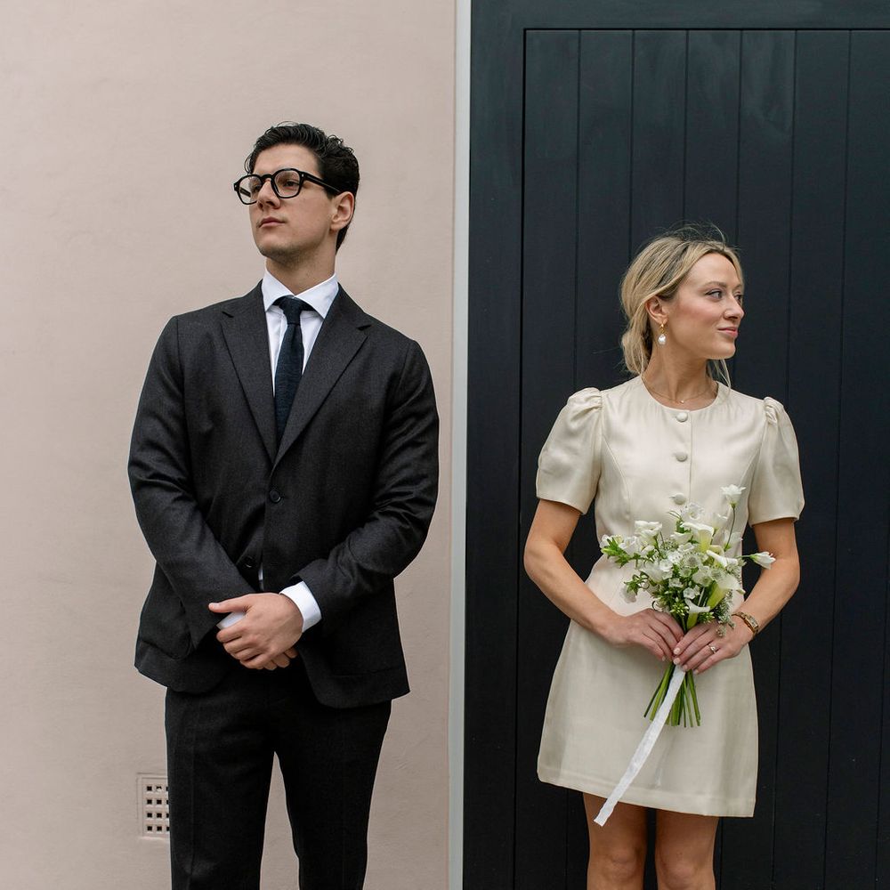 Bride in a short cream dress holding a bouquet stands beside a groom in a charcoal suit against a pink wall