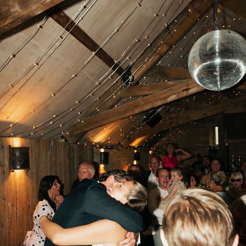 Bride and groom embrace on the dance floor beneath a mirrorball and fairy lights in a barn
