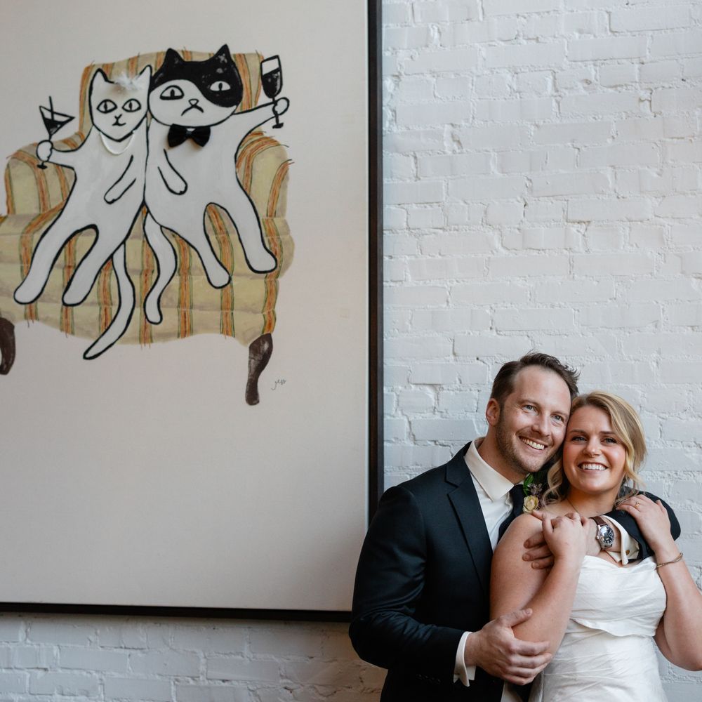 Smiling bride and groom embrace beside a quirky cat artwork on a white brick wall