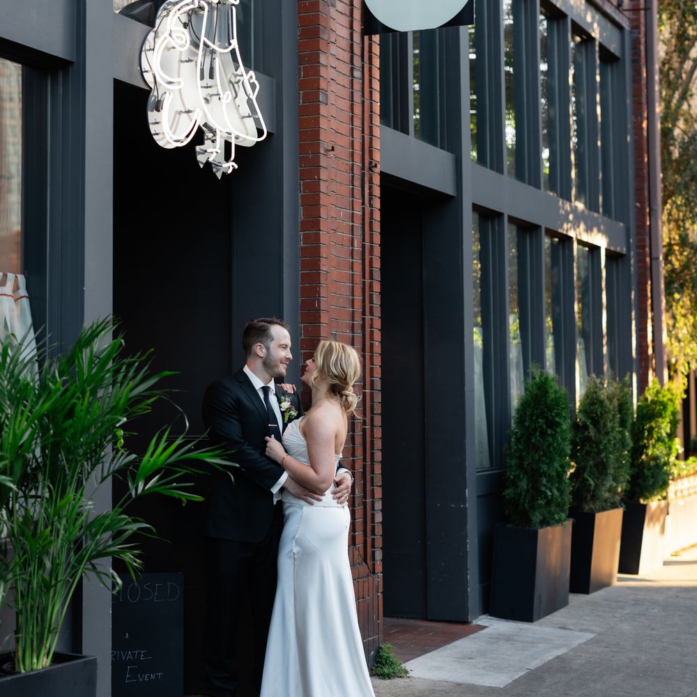 Bride and groom embracing on a city pavement outside a red-brick bistro with a neon sign