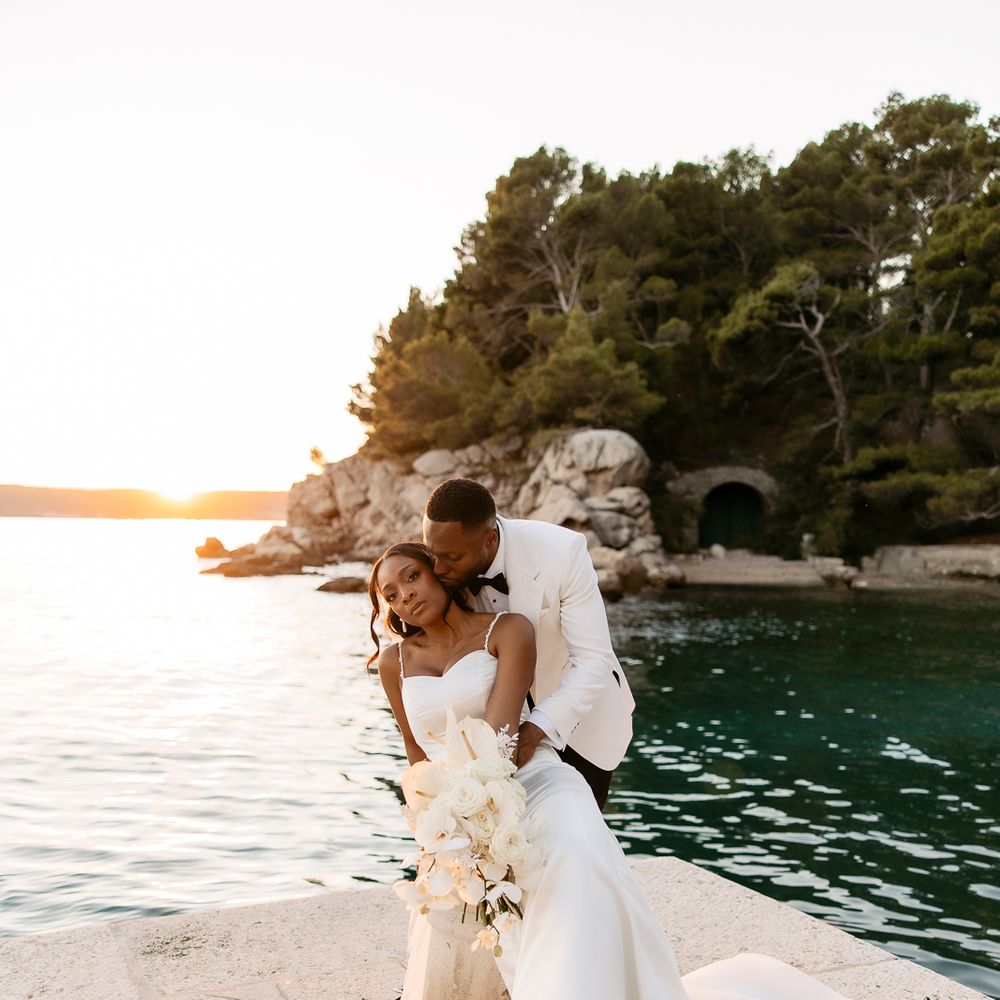 Bride and groom embrace on a stone waterside terrace at sunset with white floral styling