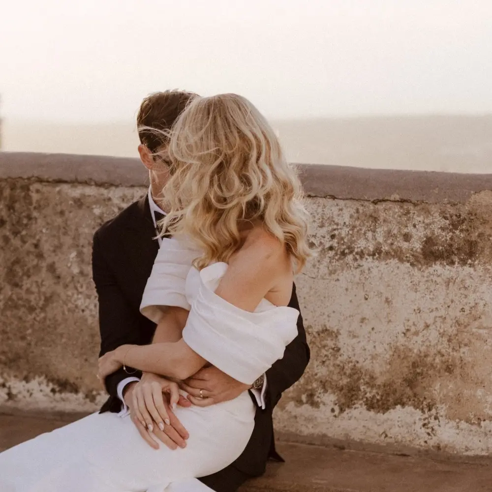 Bride in off-the-shoulder gown embraces groom on a weathered stone terrace at sunset