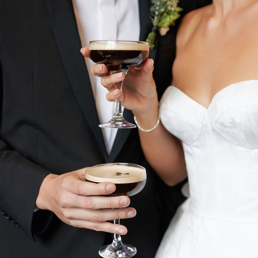 Bride and groom holding espresso martinis in coupe glasses while sharing a close embrace