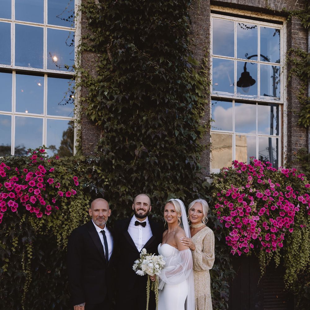 Bride and groom pose with two guests beside an ivy-clad wall bursting with pink blooms