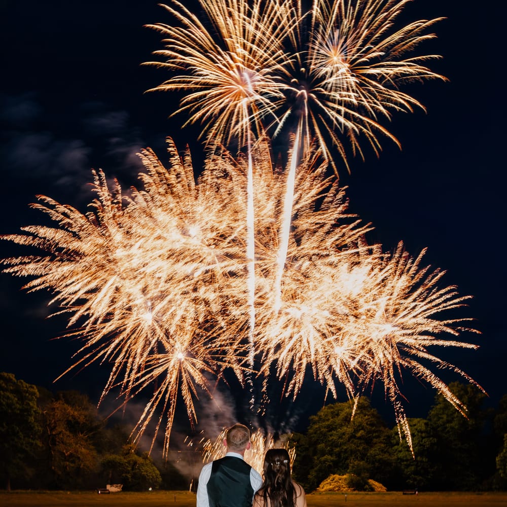 Bride and groom in kilt watch golden fireworks burst over a dark lawn at night