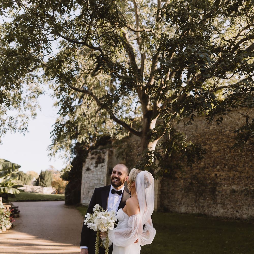 Bride in off-shoulder gown and groom in black tuxedo share a moment on a tree-lined garden path