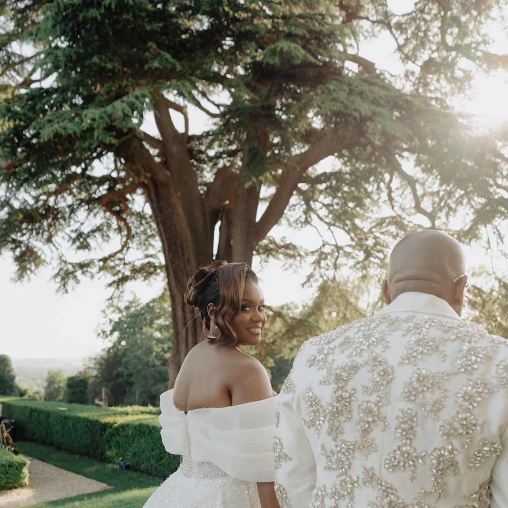 Bride in off-shoulder gown glances back beside groom in embellished jacket in a garden