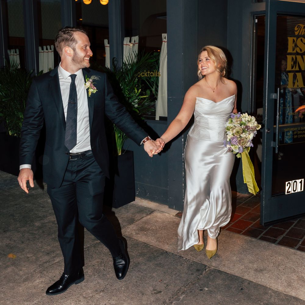 Bride in silk slip gown and groom in navy suit holding hands outside a city venue at night
