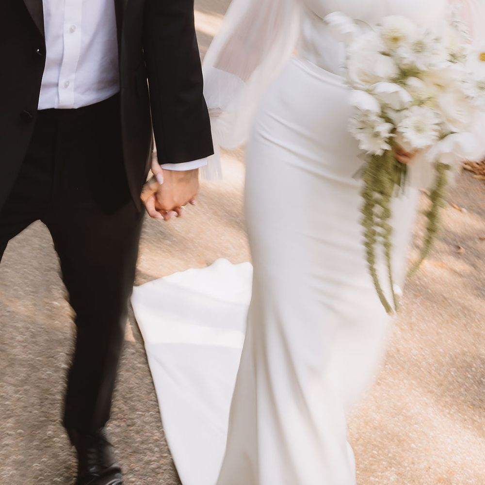 Bride in off-shoulder gown holding white bouquet walks hand in hand with groom in black tuxedo.