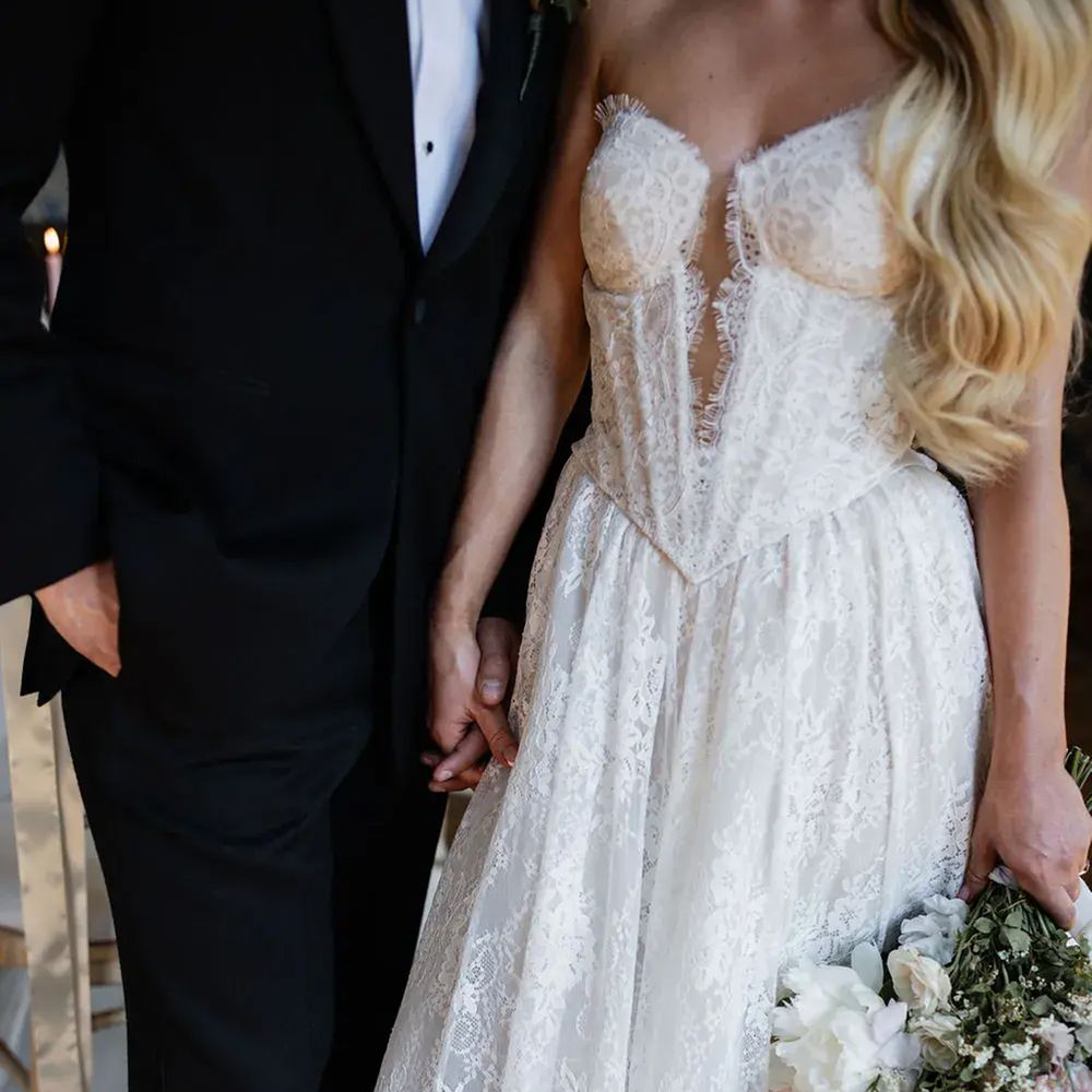 Bride in a strapless lace corset gown holding hands with groom in black tie, clutching a soft bouquet