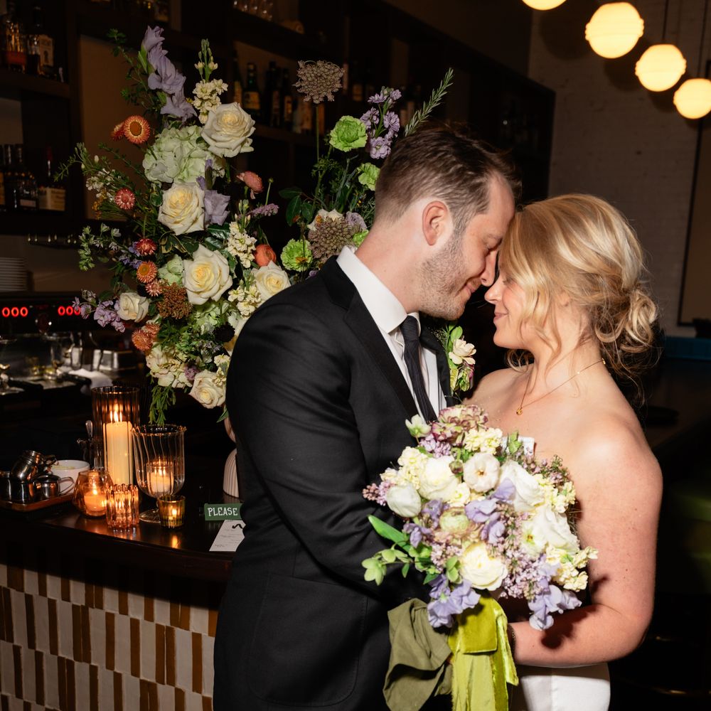 Bride and groom touch foreheads by a candlelit bar, bride holding a bouquet with green ribbon