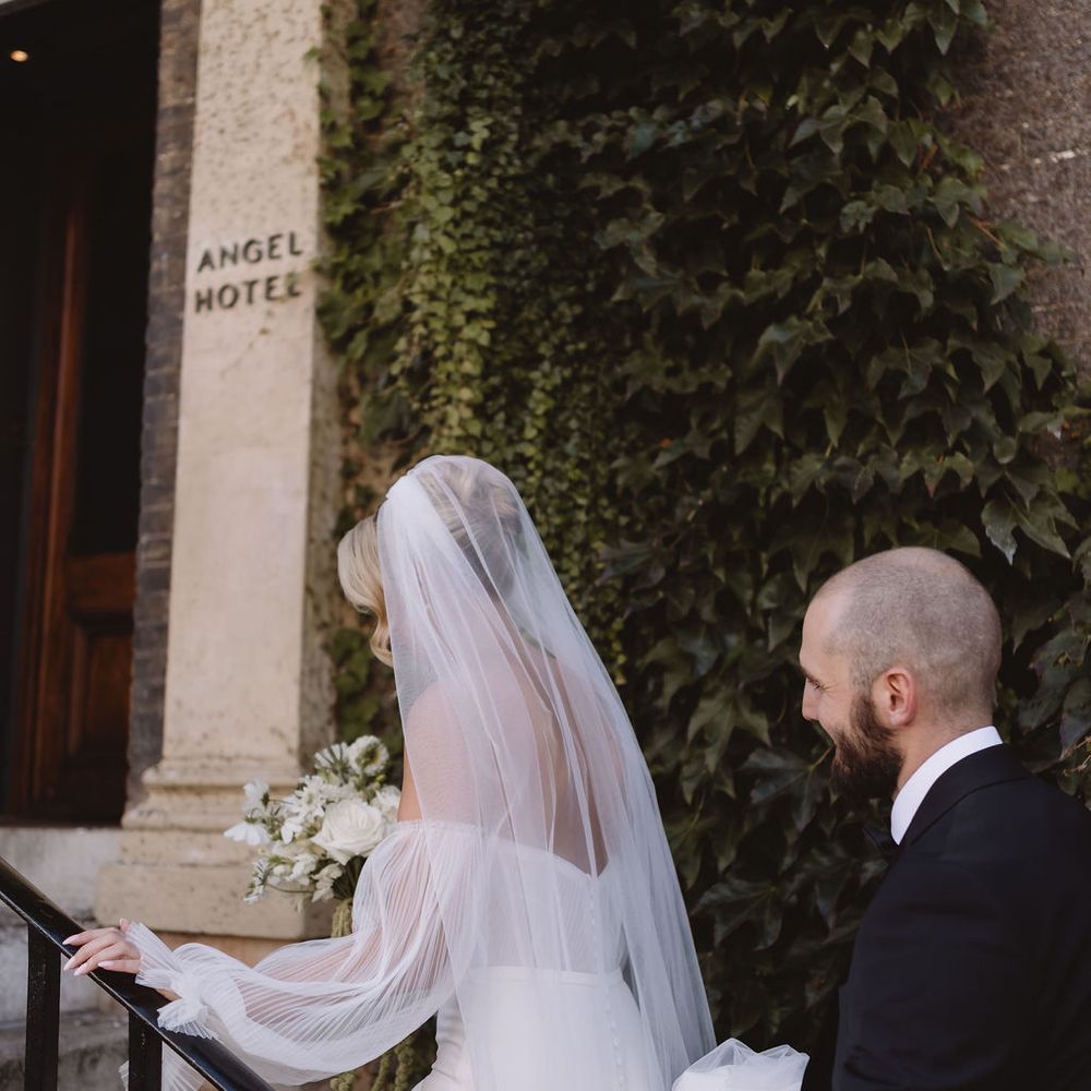 Bride in off-shoulder gown and veil ascends ivy-clad steps beside groom in black tie