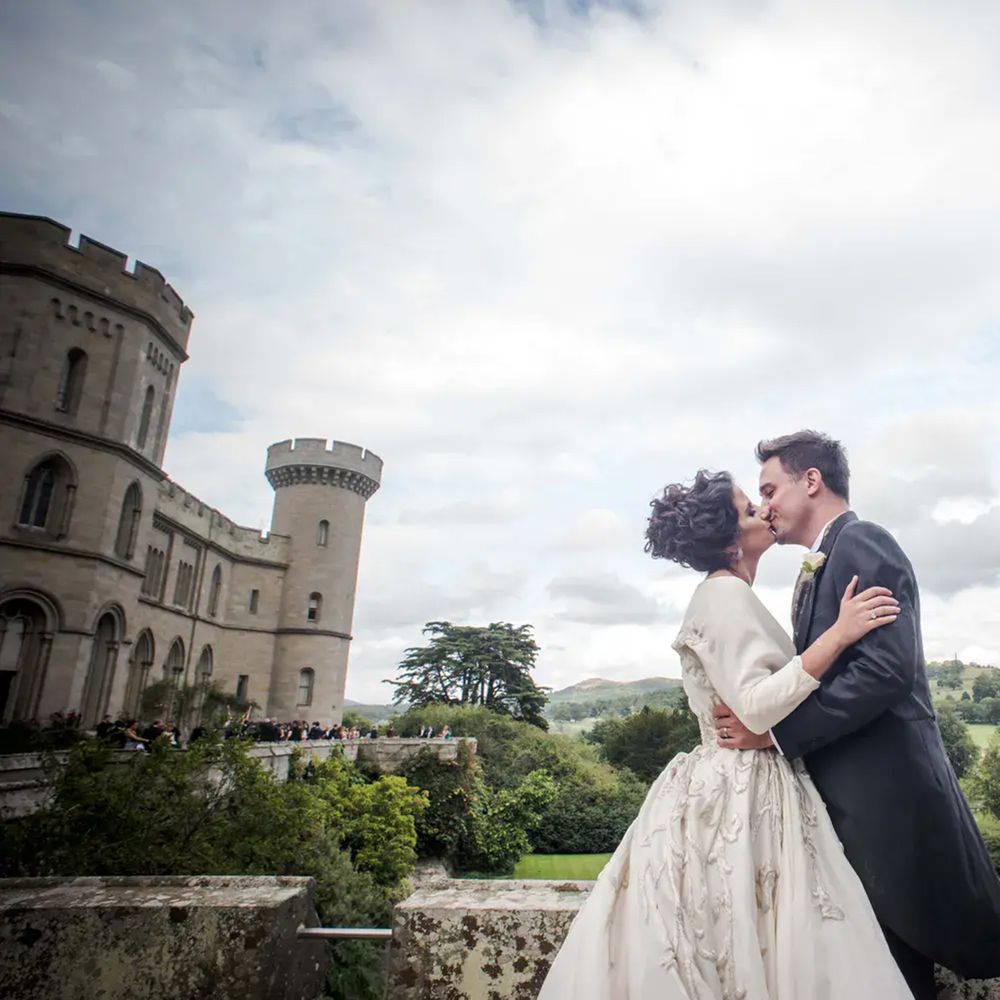 Bride and groom kiss on a castle terrace with turrets and rolling countryside behind them