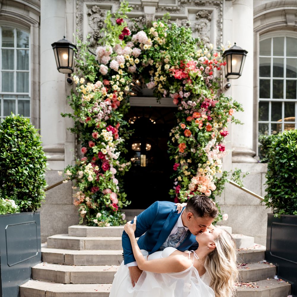 Groom in blue suit dips bride for a kiss beneath a floral archway on stone steps