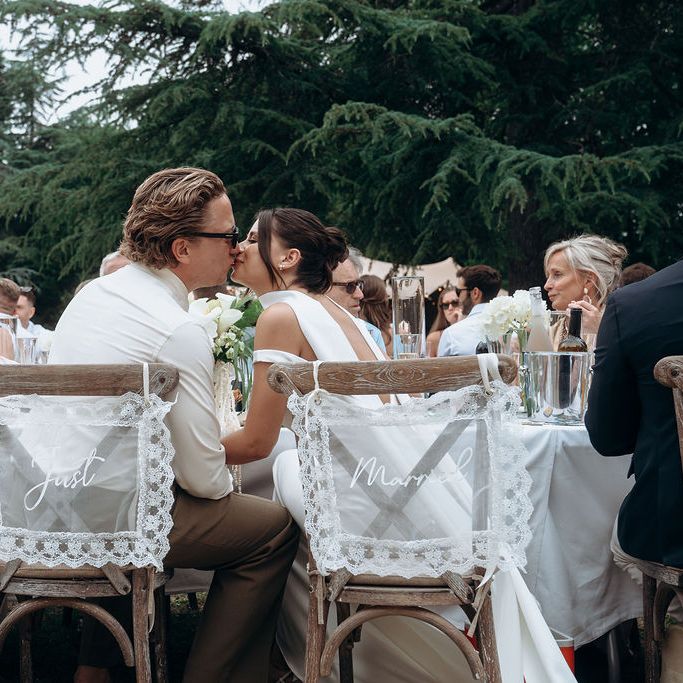Bride and groom kiss at outdoor reception with lace 'Just Married' chair signs
