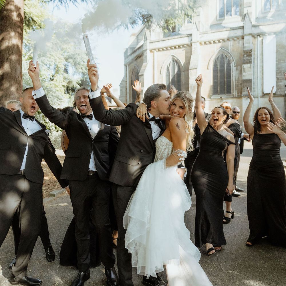 Bride and groom kiss as their wedding party cheers with white smoke bombs outside a stone chapel