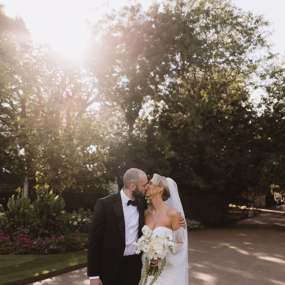 Bride and groom share a kiss on a sunlit garden pathway lined with roses and greenery