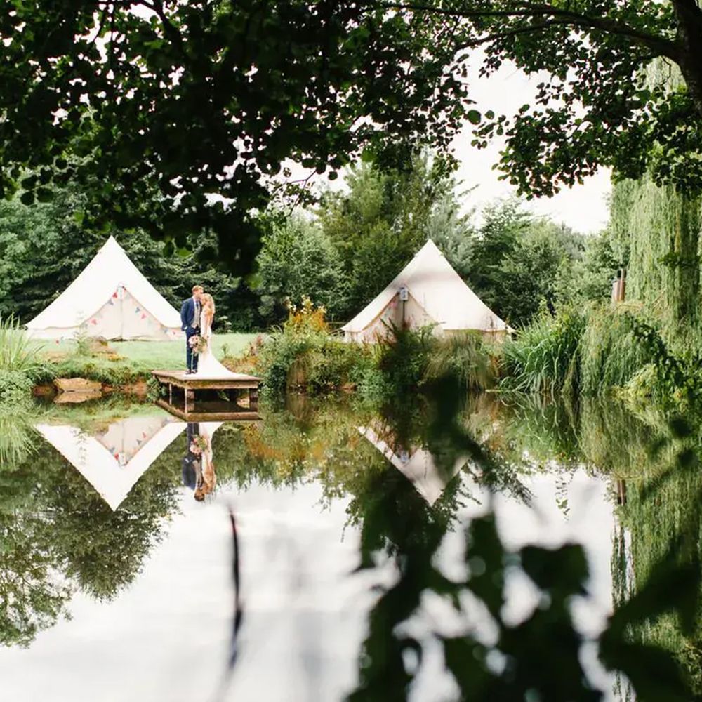 Bride and groom embrace on a wooden jetty by a lake with bell tents behind