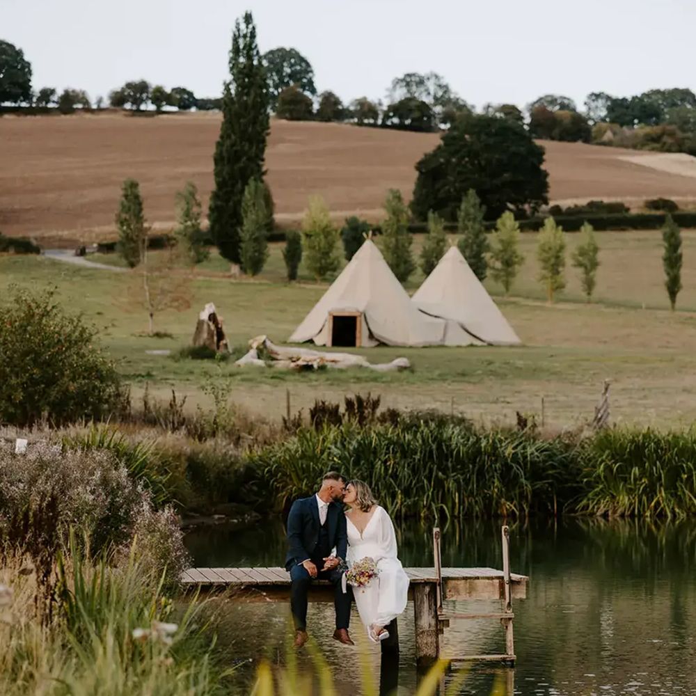 Bride and groom sharing a kiss on a lakeside jetty with tipis in the countryside behind