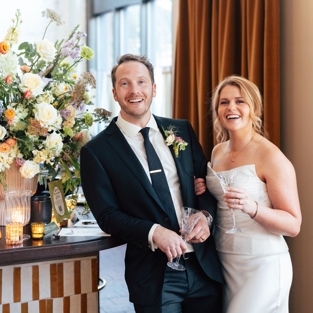 Bride and groom laughing together holding champagne glasses beside a floral arrangement