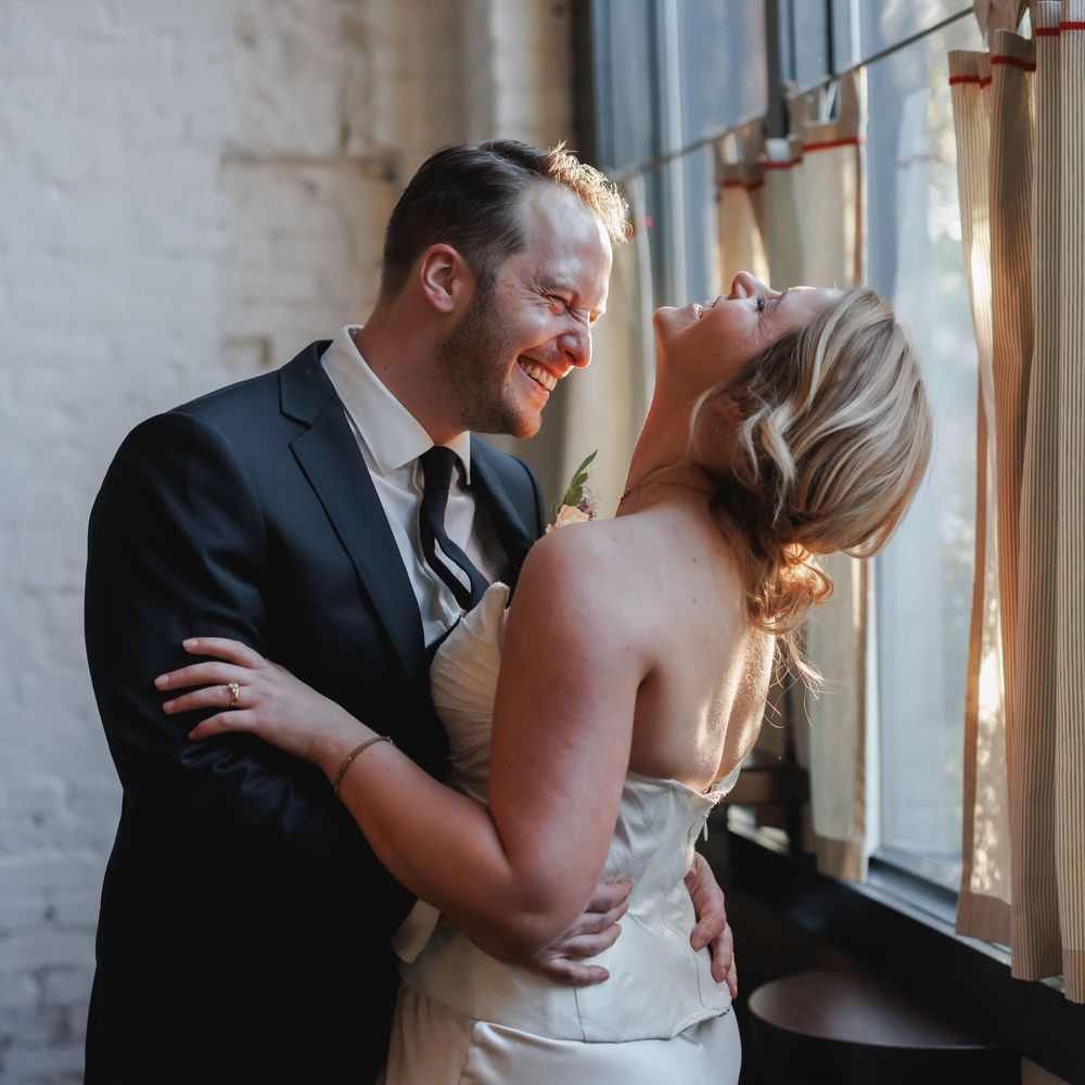 Bride and groom laughing together in an embrace beside a sunlit window with exposed brick walls