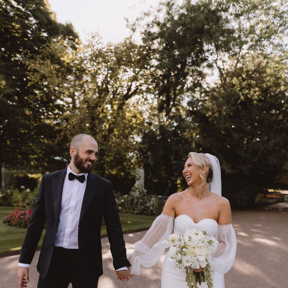 Laughing bride and groom holding hands as they walk along a tree-lined garden path