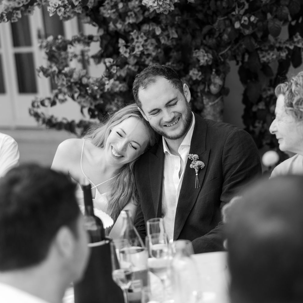 Bride resting her head on the groom's shoulder as they laugh together at an outdoor reception