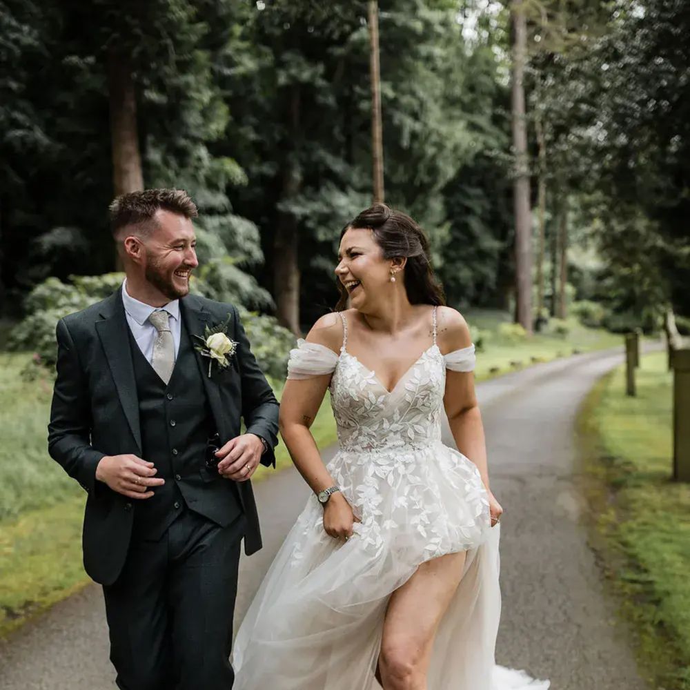 Laughing bride and groom walking along a woodland path lined with tall trees