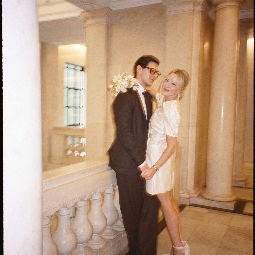 Bride in short white dress leaning on groom beside marble columns and balustrade