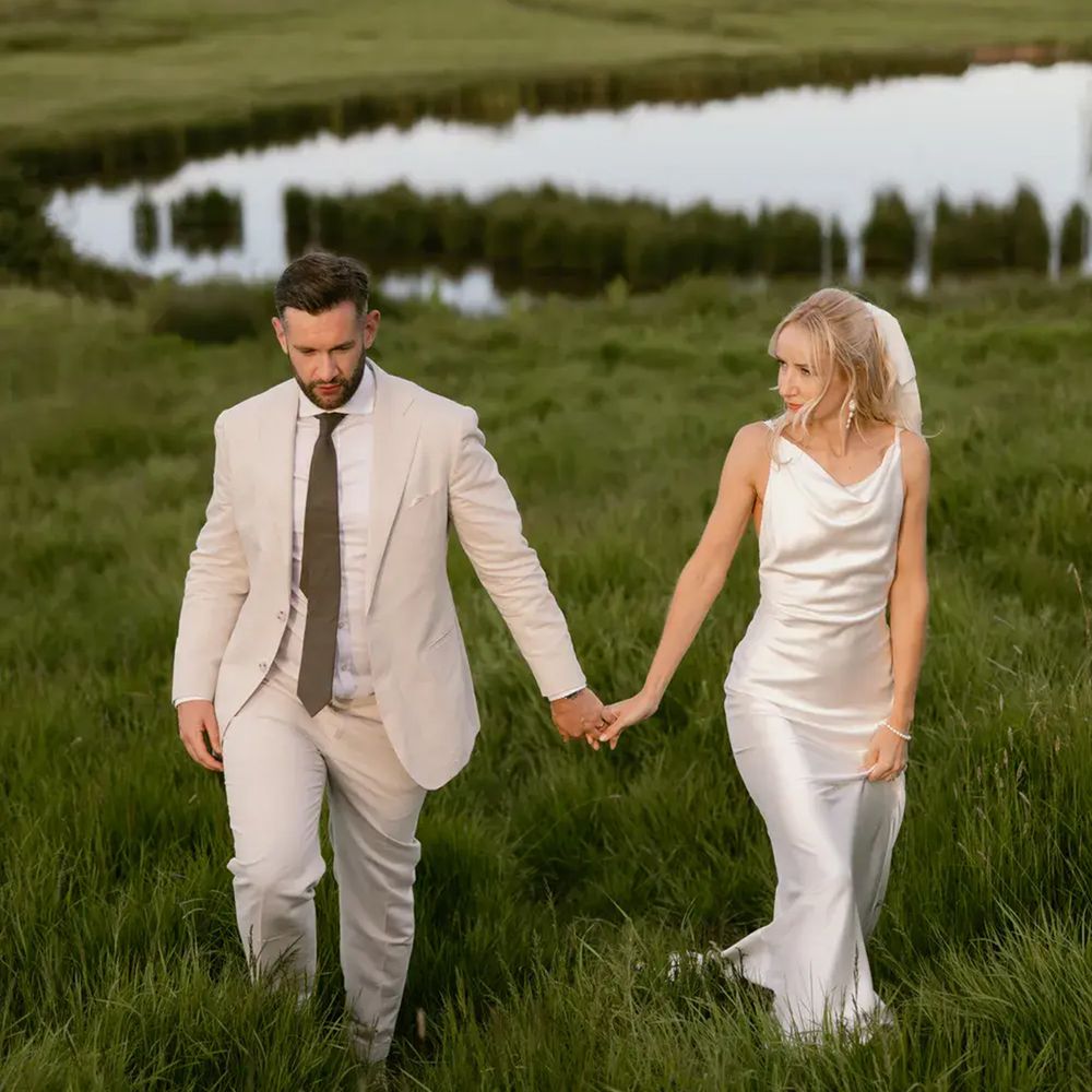 Bride and groom holding hands walking through long grass beside a still marshland pool at dusk